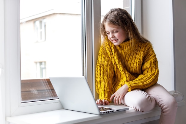 Una hermosa niña con laptop en casa en la ventana mira la pantalla y sonríe