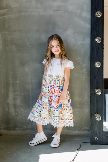 Hermosa niña linda en una blusa rosa brillante y una falda azul en una pared gris de pie cerca del espejo.