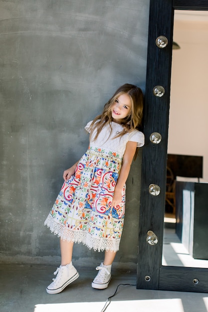 Hermosa niña linda en una blusa rosa brillante y una falda azul en una pared gris de pie cerca del espejo.