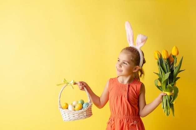 Hermosa niña linda con orejas de conejo de Pascua sostiene una canasta con huevos en un fondo amarillo