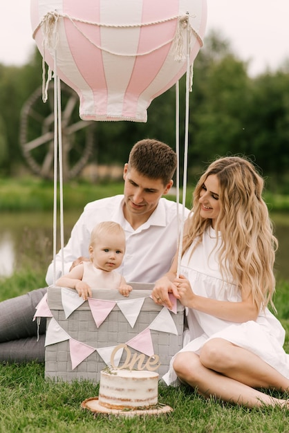 Una hermosa niña con mamá y papá en una canasta con un globo y un pastel celebra su primer cumpleaños