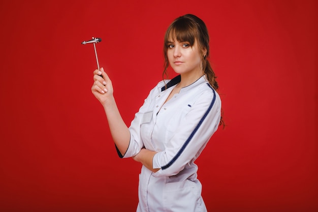 Una hermosa niña médico sostiene un martillo de reflejos y sonríe a la cámara aislada sobre un fondo rojo.