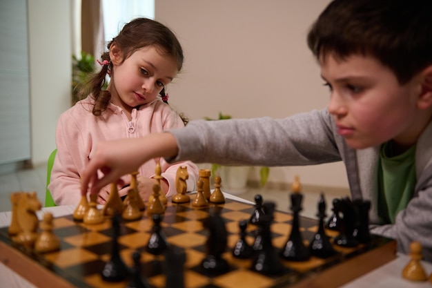 Hermosa niña mirando a su hermano moviendo su pieza de ajedrez sobre el tablero de ajedrez, haciendo su movimiento mientras jugaban ajedrez juntos en casa
