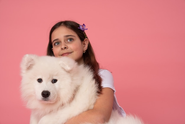 Hermosa niña morena con flor de primavera en el pelo abrazando cachorro samoyedo blanco y mirando hacia arriba en rosa