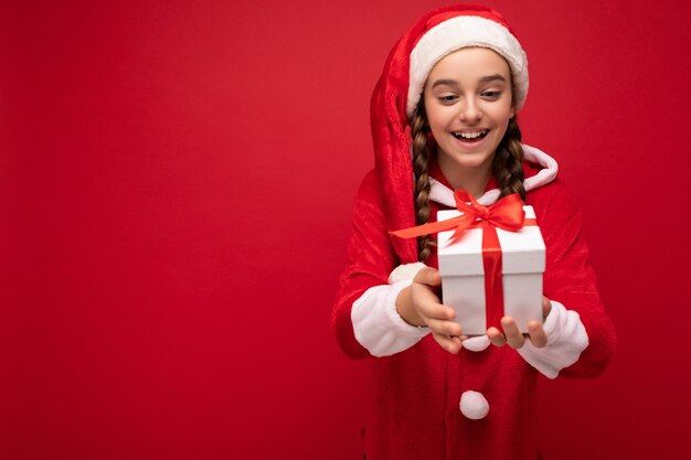 Hermosa niña morena sonriente feliz aislada sobre pared roja vistiendo traje de santa claus con caja de regalo blanca con cinta roja. Espacio vacio