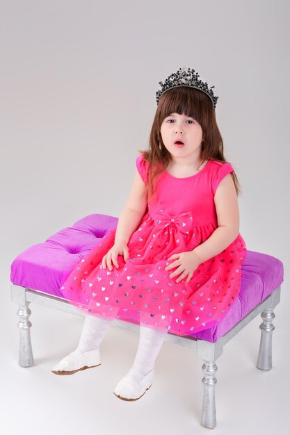 Hermosa niña morena vestida de princesa rosa con corona sentada en una silla rosa sobre fondo gris. lindo bebé