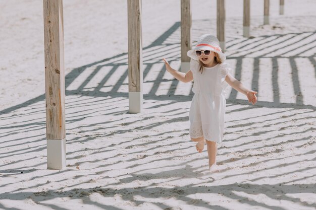 Hermosa niña morena con vestido blanco y sombrero jugando en la playa en verano