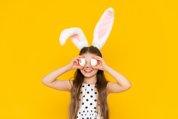 Una hermosa niña con orejas de conejo de piel en la cabeza sostiene huevos de pollo en sus manos para pintarlos para las vacaciones de Pascua
