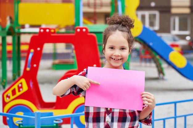 Una hermosa niña en un patio de recreo con una hoja de papel rosa vacía