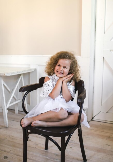 Una hermosa niña de pelo rizado sentada en medio de una habitación grande en una silla