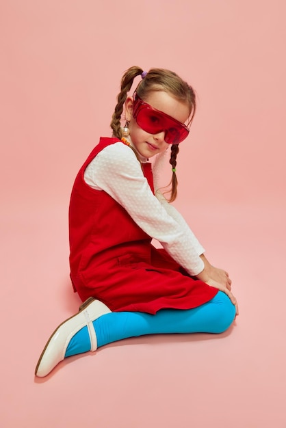 Hermosa niña pequeña con estilo en vestido rojo medias azules y gafas rojas posando sobre rosa