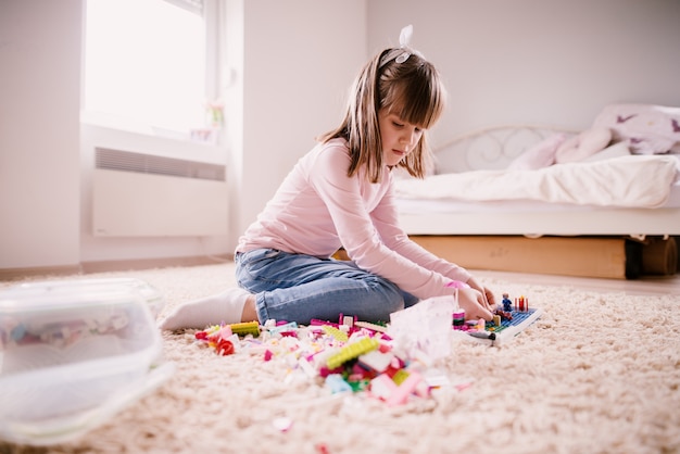 Hermosa niña pequeña pensativa sentada en la alfombra de su habitación luminosa y jugando con juguetes de plástico.