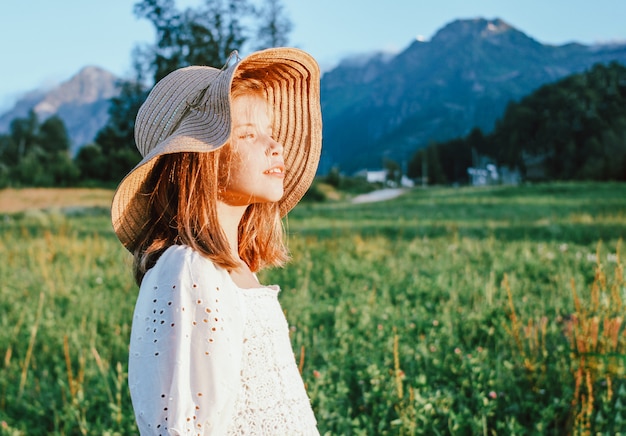 Hermosa niña preadolescente romántica con sombrero de paja sobre fondo de hermosas casas en la montaña, escena rural al atardecer