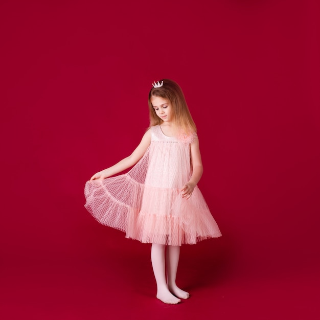 Hermosa niña princesa bailando en un lujoso vestido rosa y plateado aislado en la pared roja Cara divertida, diferentes emociones