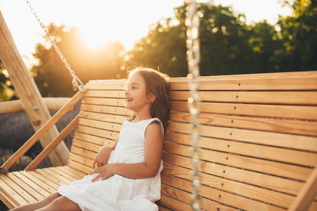 Hermosa niña riendo mientras se divierte en un columpio de madera al aire libre contra la puesta de sol.