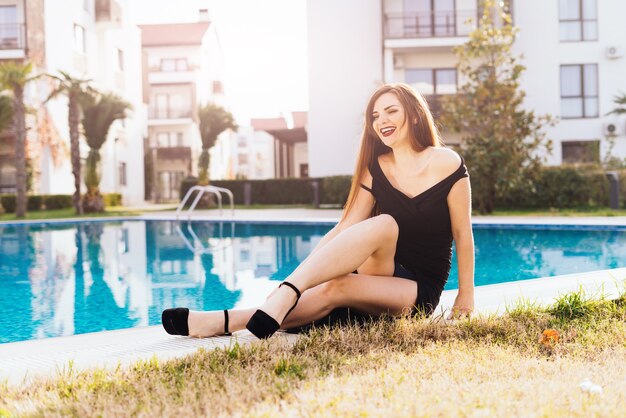 Una hermosa niña riendo con un vestido negro y zapatos se sienta junto a la piscina azul, disfruta de una vida rica y descansa