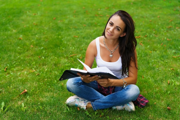 Una hermosa niña en ropa casual se sienta en la hierba en un parque universitario con un bloc de notas con un chee ...
