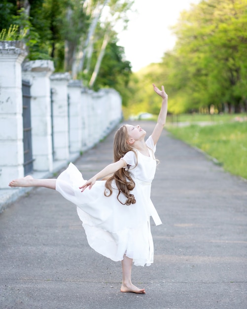 Hermosa niña rubia de años con el pelo largo con un vestido de seda blanco bailando ballet en el parque