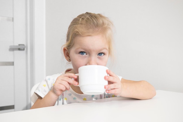 Hermosa niña rubia bebe de una taza blanca en una mesa blanca