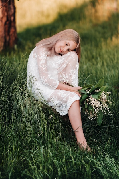 Una hermosa niña rubia de nueve años con cabello largo con un vestido largo blanco, sosteniendo un ramo de lirios del valle, sentada sobre una piedra en el parque. Verano, atardecer.