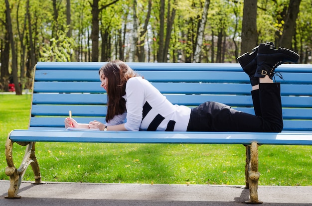 Hermosa niña de salmuera sentada en un banco, soñando y escribiendo sus planes en un cuaderno, relajándose en un parque en un soleado día de primavera