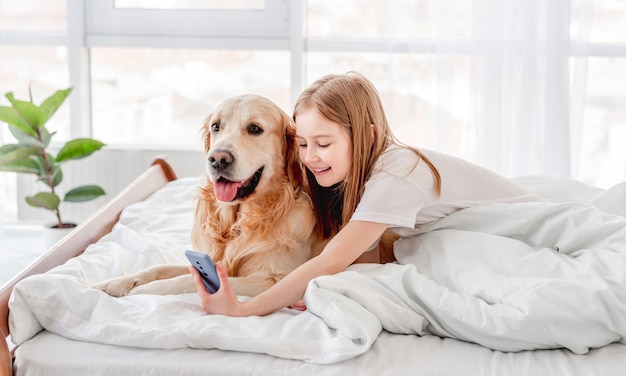 Hermosa niña sentada en la cama con perro golden retriever, haciendo selfie con smartphone y sonriendo. Niño con perro mascota y teléfono celular en el dormitorio.