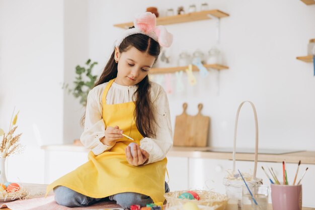 Una hermosa niña sentada en la mesa con decoraciones y pintando huevos de colores.