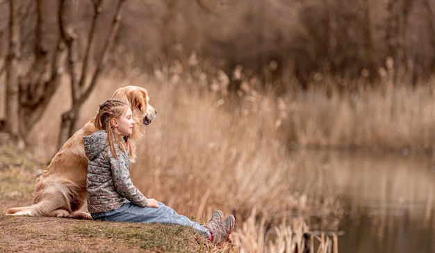 Hermosa niña sentada con perro golden retriever y mirando el río