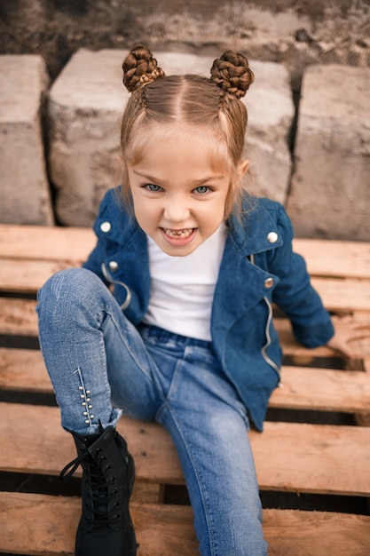 Una hermosa niña está sentada con ropa elegante en paletas de madera. niño con estilo una chaqueta azul. estilo casual, moda para niños, traje de moda, infancia feliz. | Foto Premium