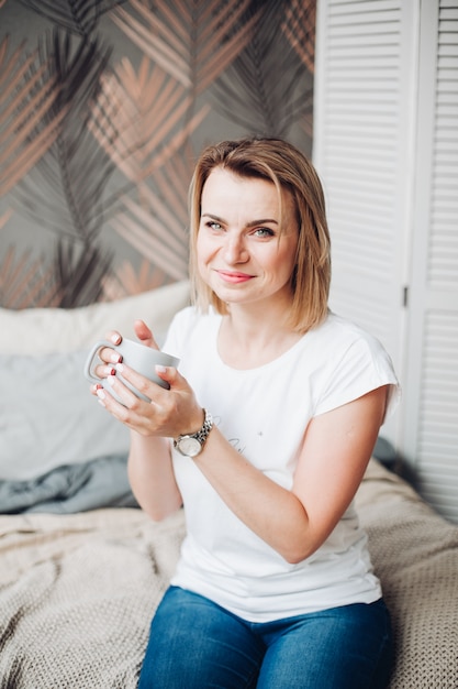 Hermosa niña sentada en su cama, disfruta de su vida y bebe un delicioso café o té