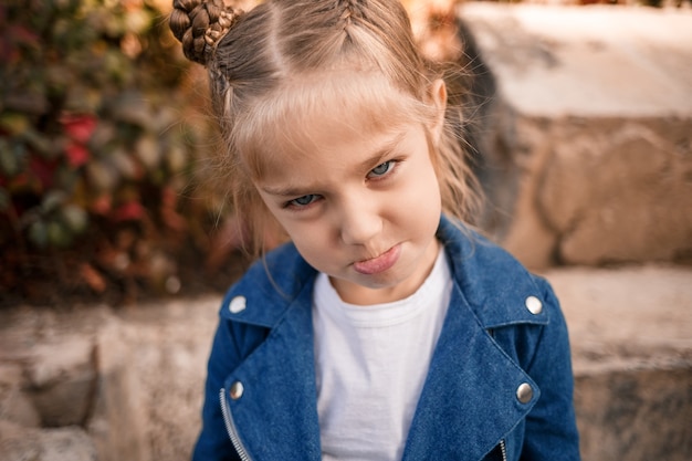 Una hermosa niña de unos siete años con una chaqueta de mezclilla mira a la cámara y hace muecas