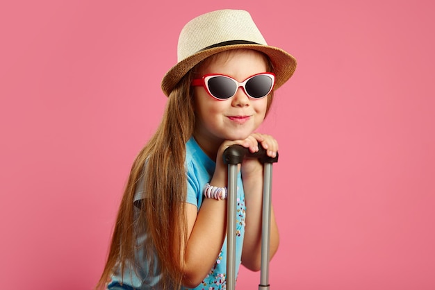 Hermosa niña de siete años con gafas de sol y sombrero de paja se apoya en el mango de la maleta