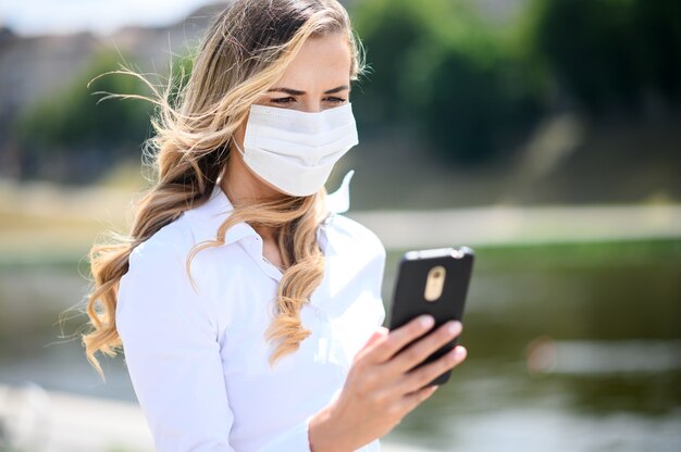 Hermosa niña sola mujer joven usando su teléfono inteligente y con una máscara, concepto de coronavirus