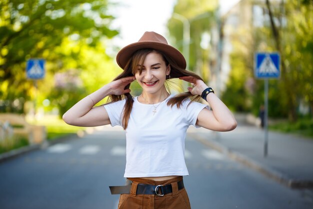 Una hermosa niña con un sombrero marrón y una camiseta blanca está caminando por la carretera en un día cálido y soleado. Hermosa mujer joven con un sombrero con una sonrisa en su rostro