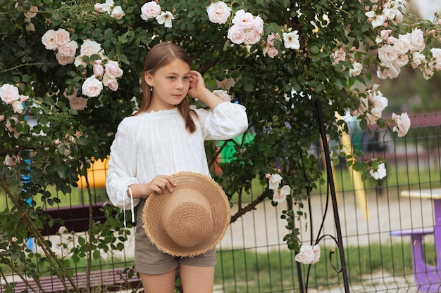 Foto hermosa niña con sombrero de paja cerca de un arbusto de rosas blancas