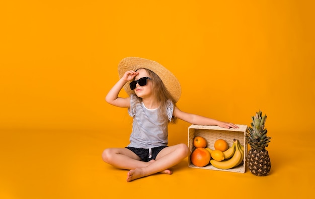 Una hermosa niña con un sombrero de paja y gafas de sol se sienta con una canasta de frutas sobre una superficie amarilla y mira hacia otro lado