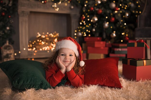 Hermosa niña con un sombrero rojo de Navidad se encuentra junto a la chimenea y el árbol de Navidad