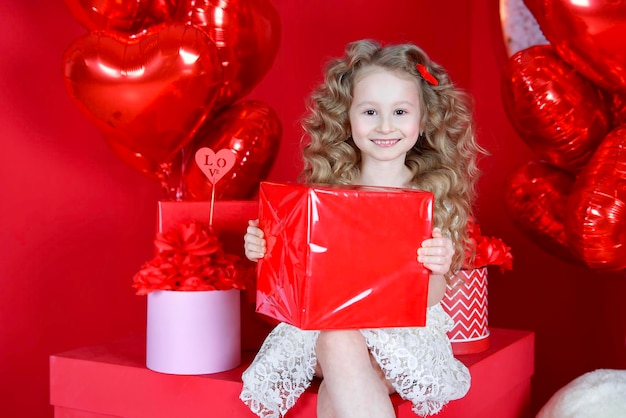 Hermosa niña sonríe sobre un fondo rojo con un globo en forma de corazón y una caja de regalo en sus manos El concepto de regalos de amor Día de San Valentín 14 de febrero 8 de marzo día de la madre