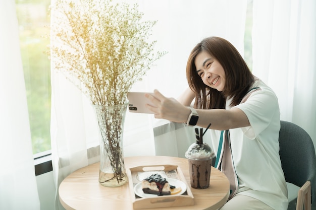 Hermosa niña sonriendo con teléfono móvil selfie en la cafetería, niña come café y pastel