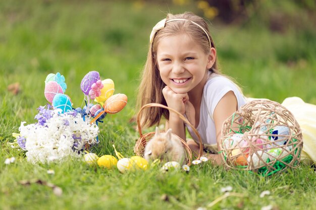 Hermosa niña sonriente acostada en la hierba junto a un lindo conejito, huevos de Pascua y flores en las vacaciones de primavera. Mirando a la cámara.