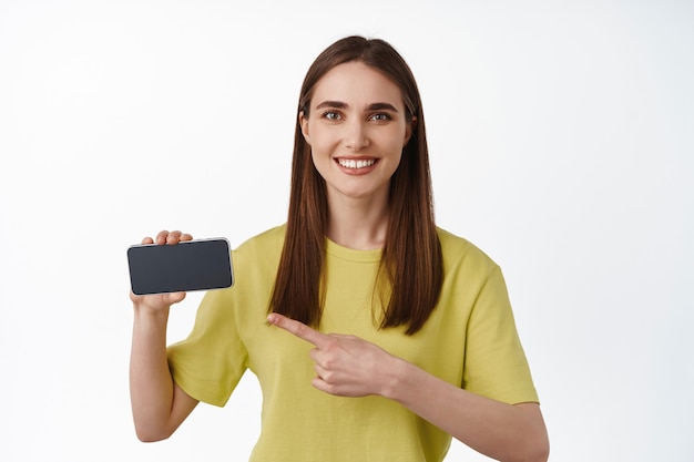 Hermosa niña sonriente apuntando a la pantalla horizontal del teléfono inteligente, mostrando el anuncio en el teléfono, interfaz de la aplicación, de pie en una camiseta en blanco
