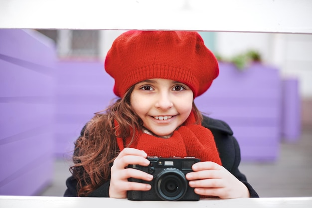 Hermosa niña sonriente en una boina roja y un pañuelo con una cámara en una calle de la ciudad. Foto de alta calidad