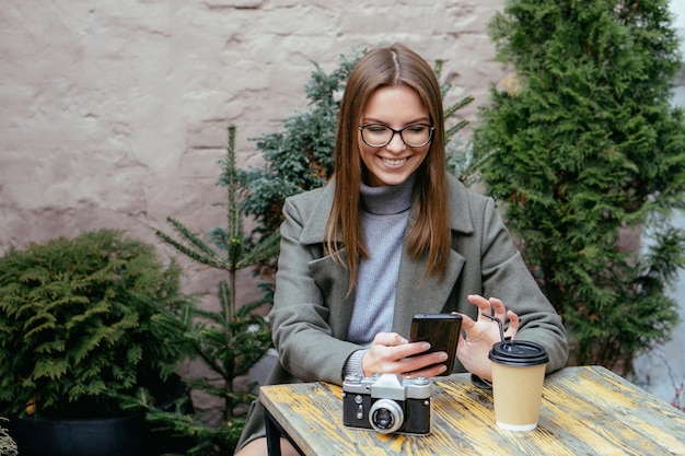 Hermosa niña sonriente con gafas sentado en la cafetería con smartphone en manos y charlando