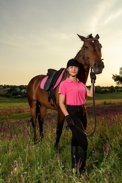 Hermosa niña sonriente jockey de pie junto a su caballo marrón con uniforme especial sobre un fondo de cielo y campo verde en una puesta de sol.