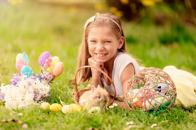 Hermosa niña sonriente con un lindo conejito en la canasta y huevos de Pascua en la hierba en las vacaciones de primavera. Mirando a la cámara.