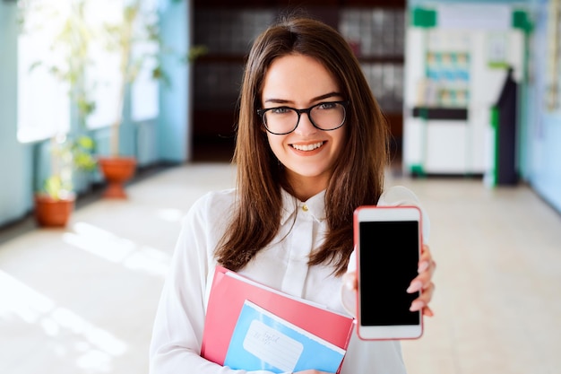 Hermosa niña sonriente con materiales de aprendizaje y un teléfono en el pasillo de la universidad.