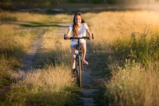 Hermosa niña sonriente montando en bicicleta en prados al atardecer