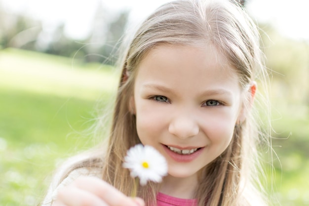 Hermosa niña sonriente con ojos azules en el parque sosteniendo una flor blanca y mirando a la cámara.