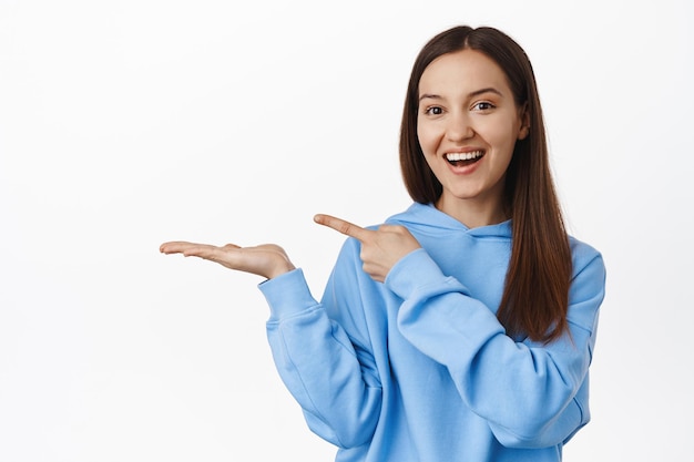 Hermosa niña sonriente señalando su mano abierta, mostrando un artículo en la palma, publicitando algo, mostrando el producto contra el fondo blanco