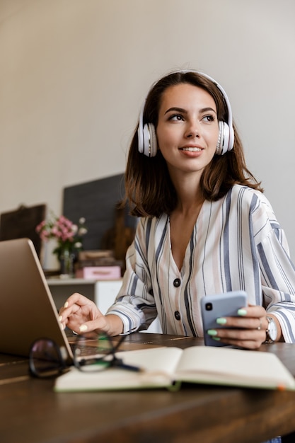 Foto hermosa niña sonriente sentada en la mesa en casa, mediante teléfono móvil